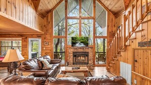 A cozy cabin interior with a stone fireplace, leather sofa, and large windows offering a forest view. - Raccoons' Rest: Kick Back in the Smokies (Pigeon Forge)