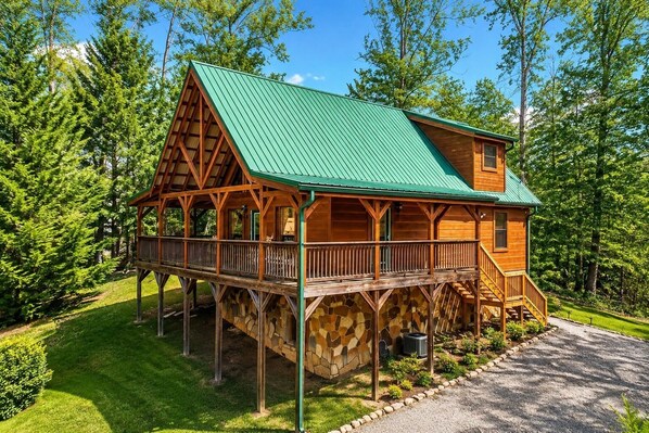 A wooden cabin with a green roof, a stone foundation, and a wooden deck.
