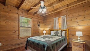 Un dormitorio de cabaña de madera con una cama, dos mesitas de noche, un ventilador de techo y una ventana con persianas.