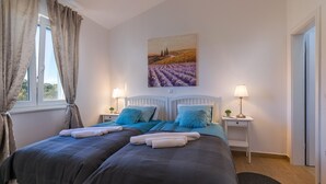 A double bed with a dark blue and gray duvet, two pillows, and a matching blanket. There are two bedside tables with lamps, a painting on the wall, and a window with curtains. - Villa Klara in Klarići (Klarići)