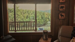 Una habitación con un ventanal grande que da a un área boscosa, una silla de madera y una pequeña mesa con una vela.