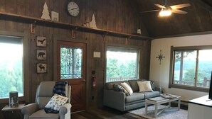 Una habitación acogedora con techo de madera, un sofá, una silla y una mesa de centro. Hay ventanas con vistas a árboles y una puerta con un ventanal.