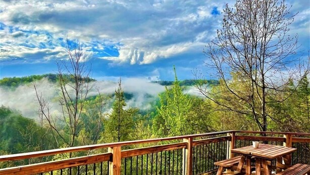 Una terraza de madera con una mesa y bancos, con vistas a un valle boscoso cubierto de niebla y un cielo despejado.