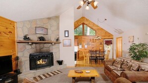 Ein gemütliches Wohnzimmer mit Kamin aus Stein, Holzmöbeln und einem Essbereich mit hohen Decken und Tageslicht.