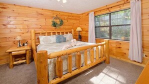 Ein Zimmer in einer Holzhütte mit einem Bett, einem Nachttisch, einem Vorhangfenster und einem Deckenventilator.
