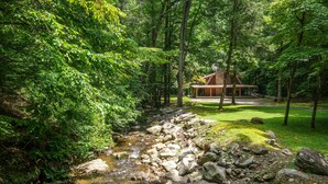 A rustic cabin surrounded by dense forest, a small stream, and a well-maintained lawn.