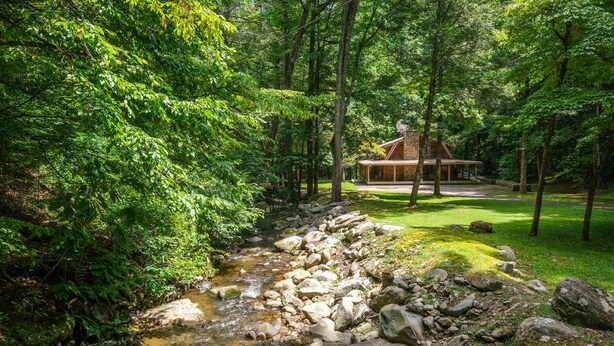 Una cabaña rústica rodeada de un bosque frondoso, un arroyo pequeño y un césped bien cuidado.