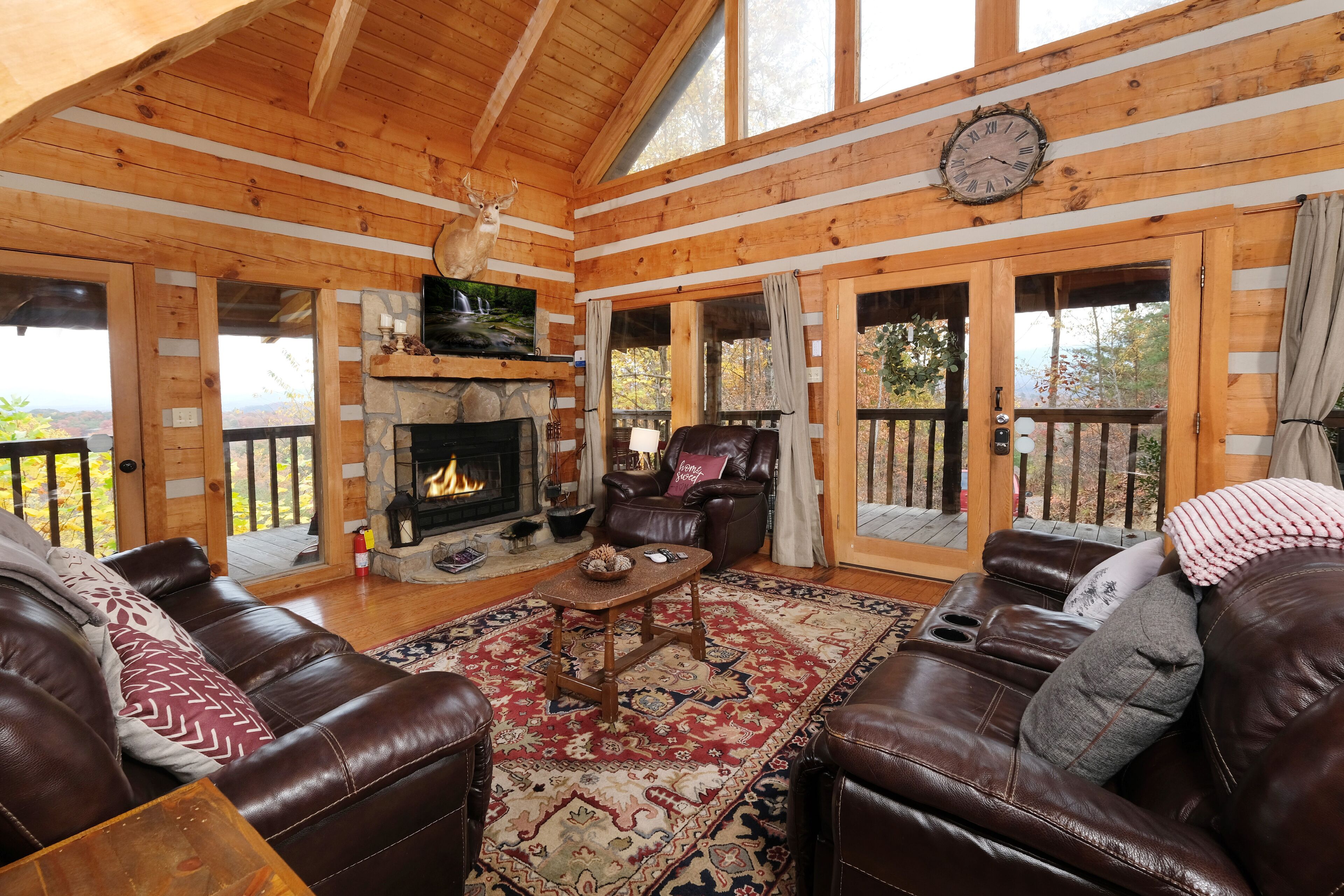 A log cabin interior with a fireplace, leather sofas, and a large window overlooking a scenic view.