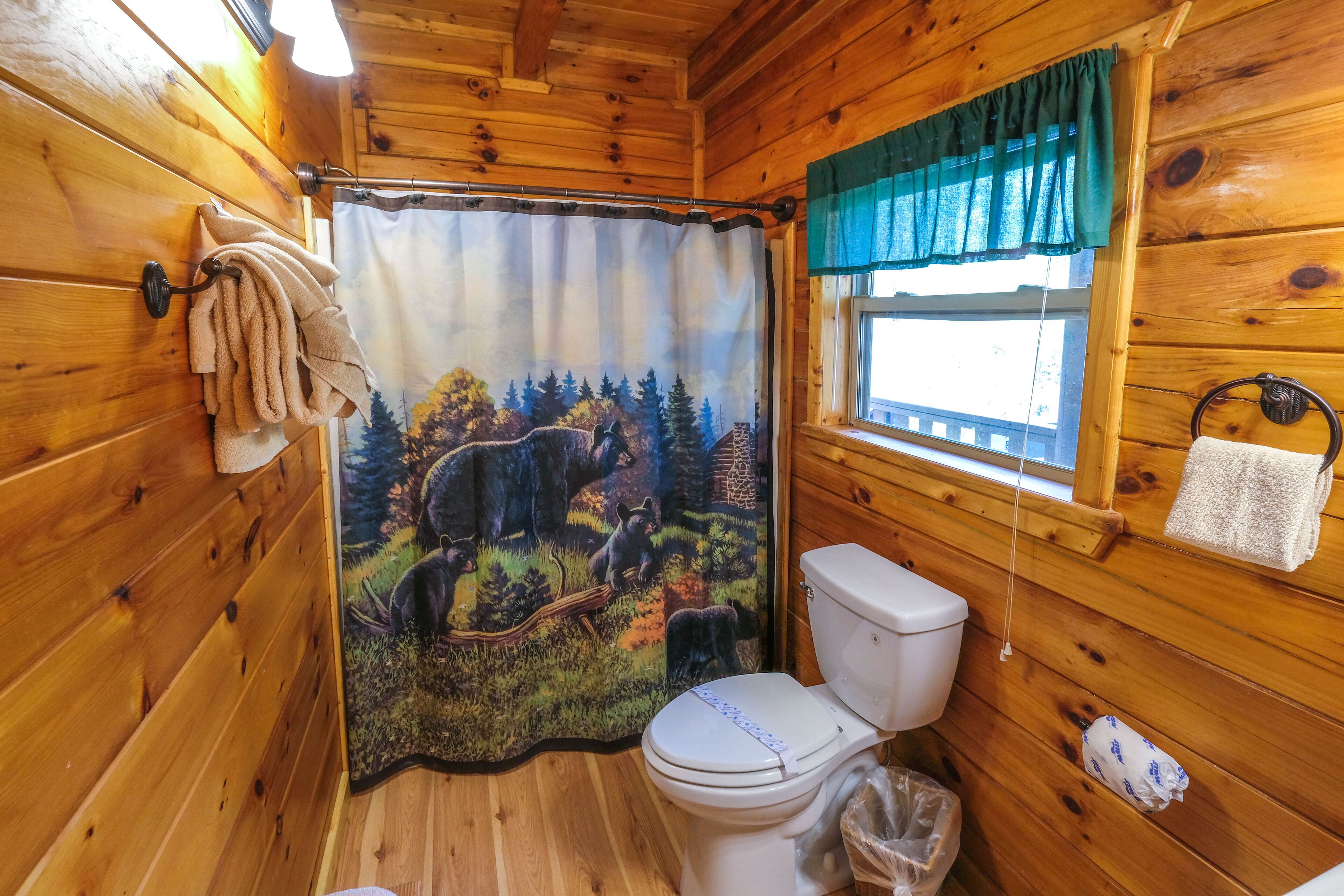 Une salle de bain rustique, avec des murs en bois, une douche ornée d’un rideau aux motifs forestiers, des toilettes et une fenêtre agrémentée d’un lambrequin vert.