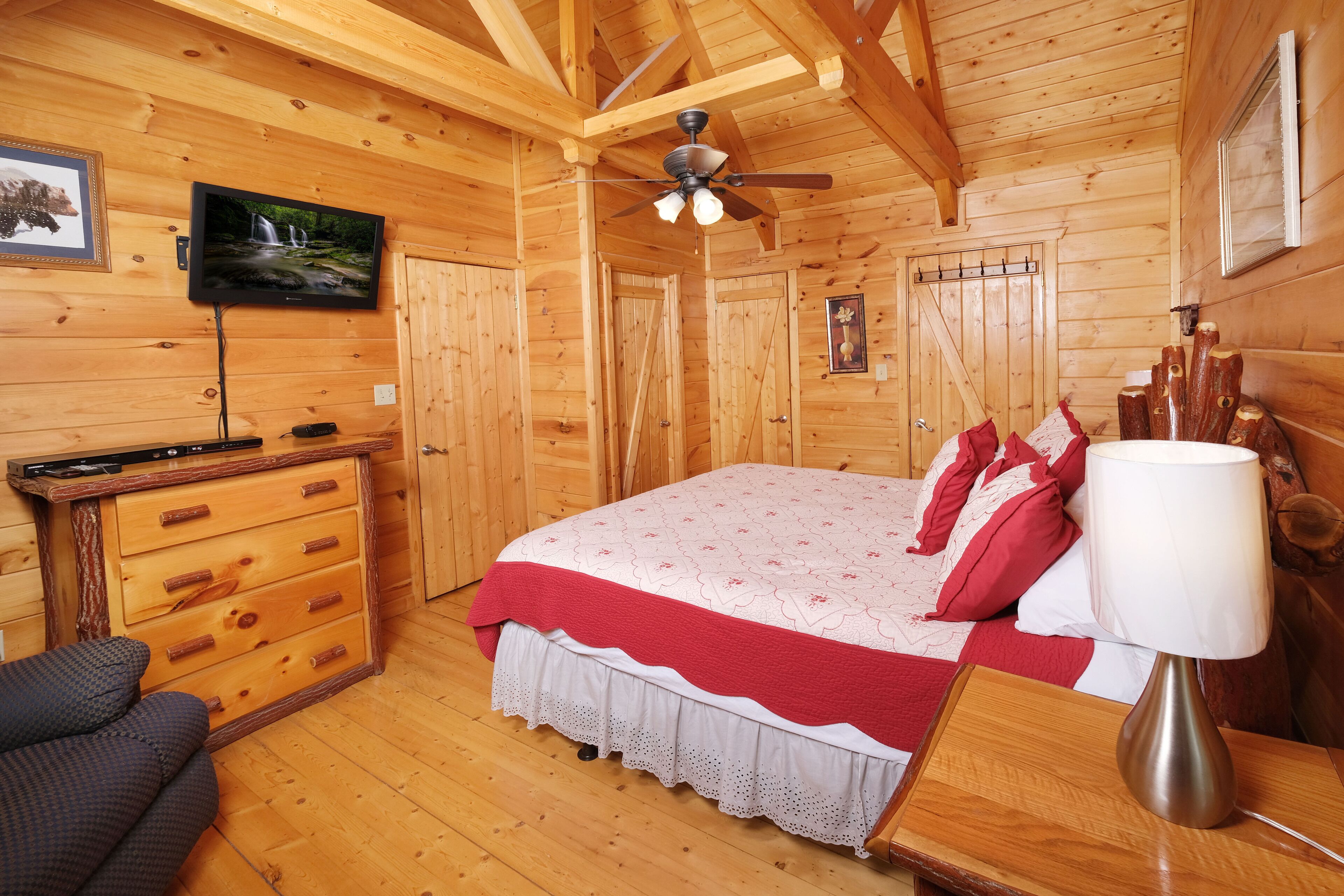 A wooden cabin bedroom with a bed, a TV, a ceiling fan, and a wooden dresser.