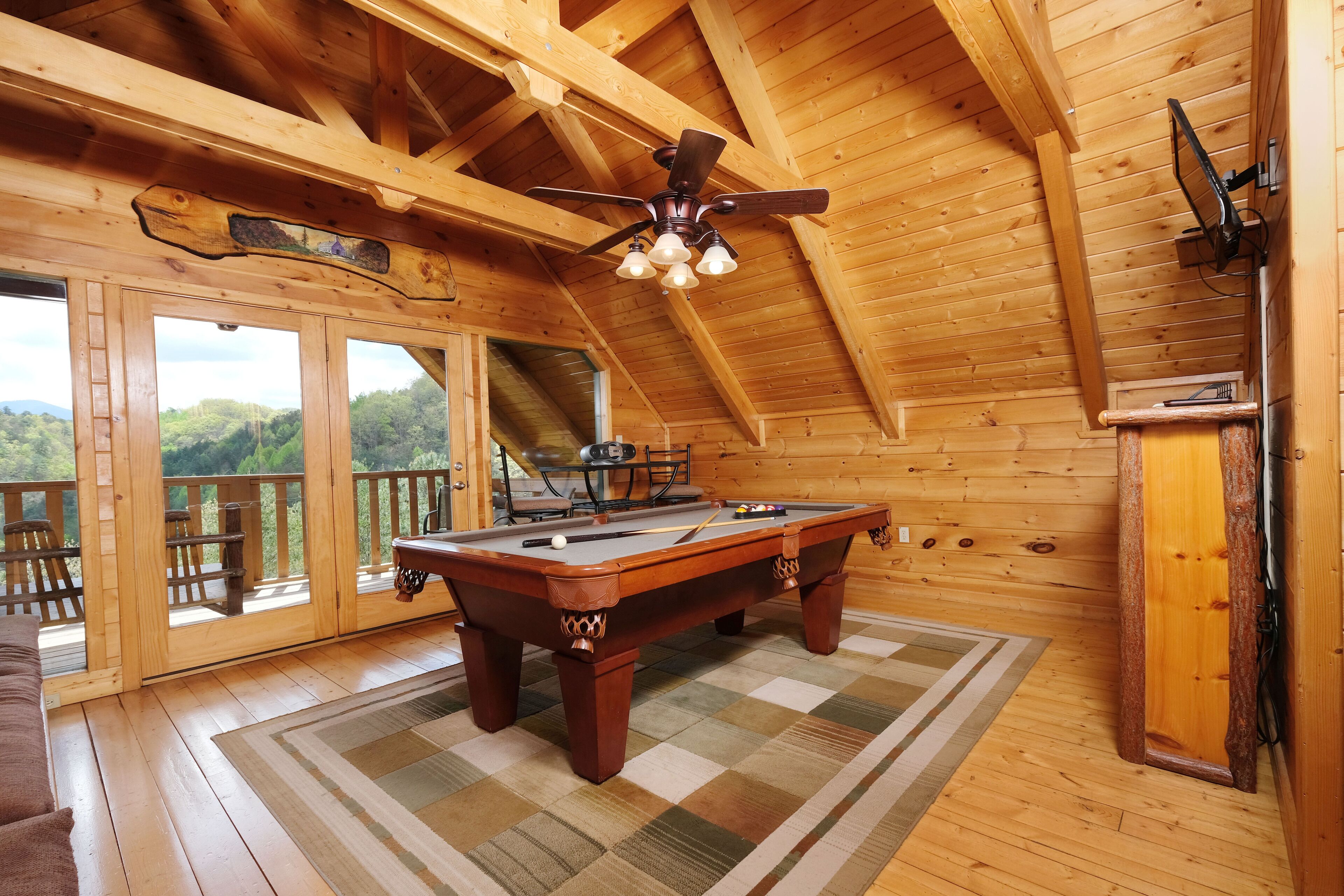 A wooden interior with a pool table, ceiling fan, and large windows overlooking a scenic view.