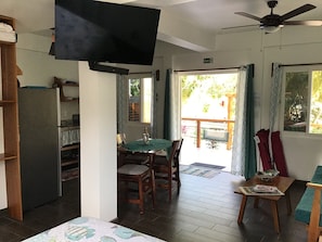 Une pièce dotée d’un téléviseur à écran plat fixé au mur, d’un ventilateur de plafond, d’un coin repas avec une table et des chaises, et offrant une vue sur l’extérieur par une porte.