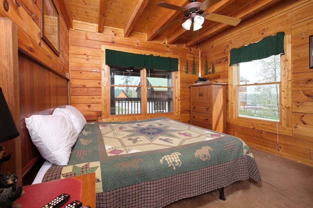 A wooden cabin bedroom with a bed, a ceiling fan, and two windows with curtains.