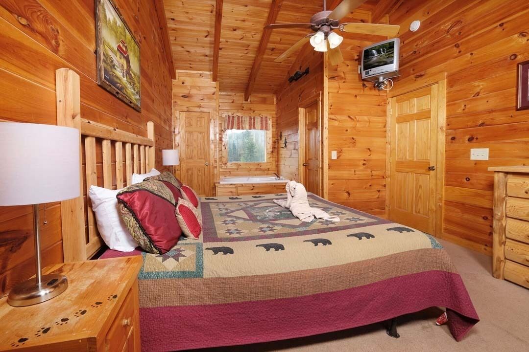 Ein Zimmer in einer Holzhütte mit einem Bett, einem Deckenventilator, einem Fernseher und einem Fenster.