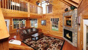 A spacious log cabin interior with a cozy living area, a stone fireplace, and a balcony with wooden railings. - Bear Ridge: Stay on the Wild Side (Pigeon Forge)
