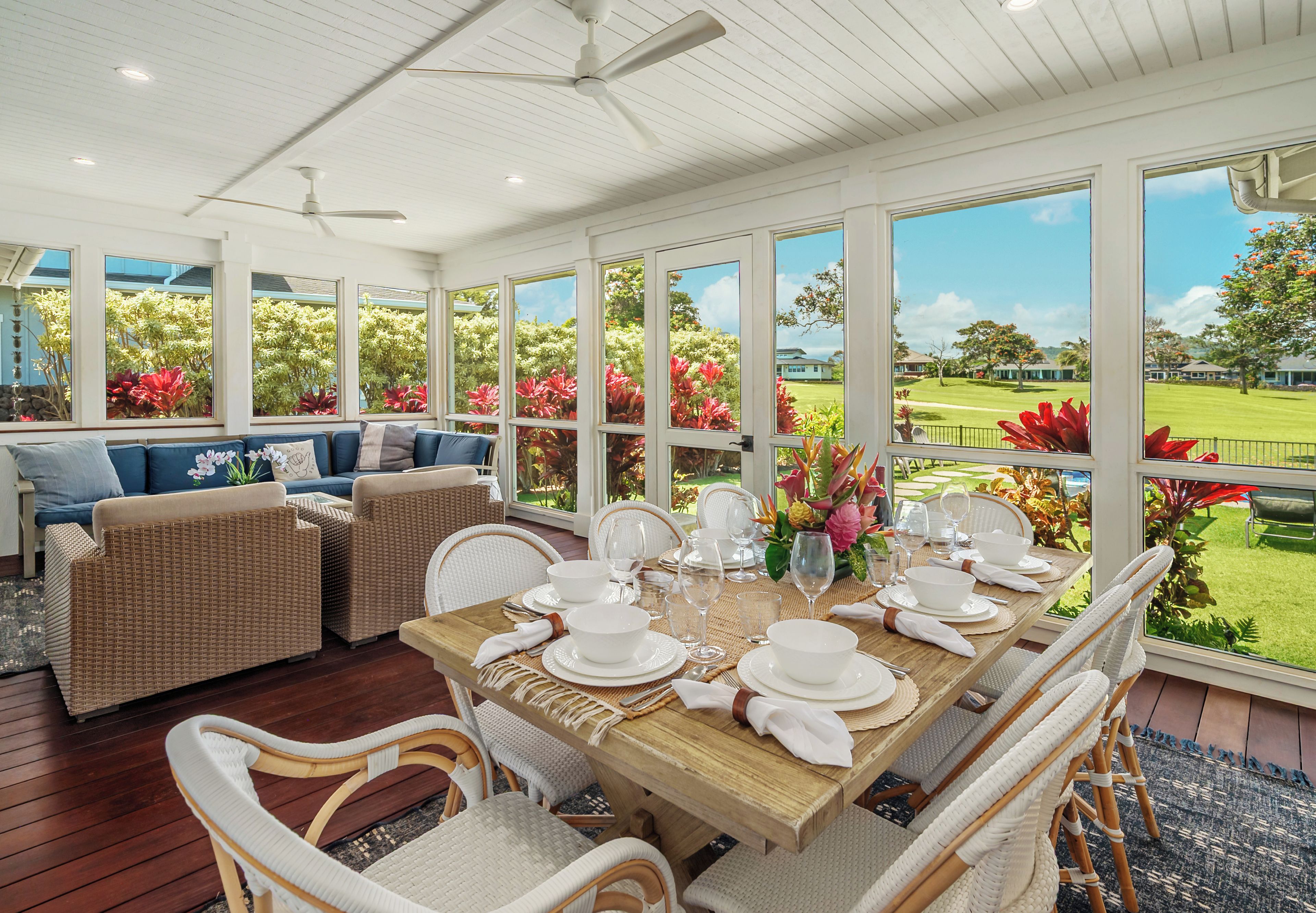 A dining area with a table set for six, surrounded by wicker chairs, with a view of a garden and a house through large windows.