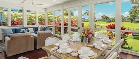 A dining area with a table set for six, surrounded by wicker chairs, with a view of a garden and a house through large windows.