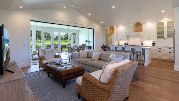 A spacious living area with a large sofa, ottoman, and wicker chairs. A kitchen with white cabinets and a dining area with a table and chairs is visible.