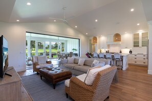 Amplia sala de estar con un sofá grande, un reposapiés y sillas de mimbre. Se ve una cocina con armarios blancos y una zona de comedor con una mesa y sillas.