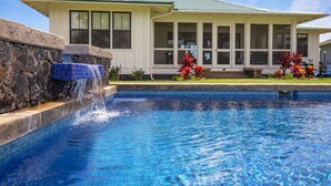 A swimming pool with a waterfall feature, a house with a green roof, and red flowers in the foreground.