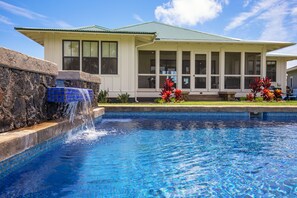 Una piscina con una cascada, una casa con techo verde y flores rojas en primer plano.