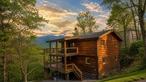 A log cabin with a balcony, a staircase, and a view of the surrounding forest and mountains.
