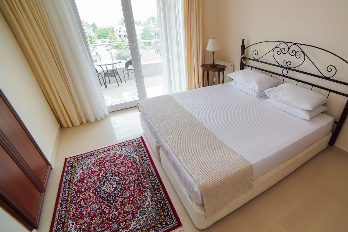 A neatly made bed with white linens and a decorative headboard, a small bedside table with a lamp, and a patterned rug on a tiled floor.