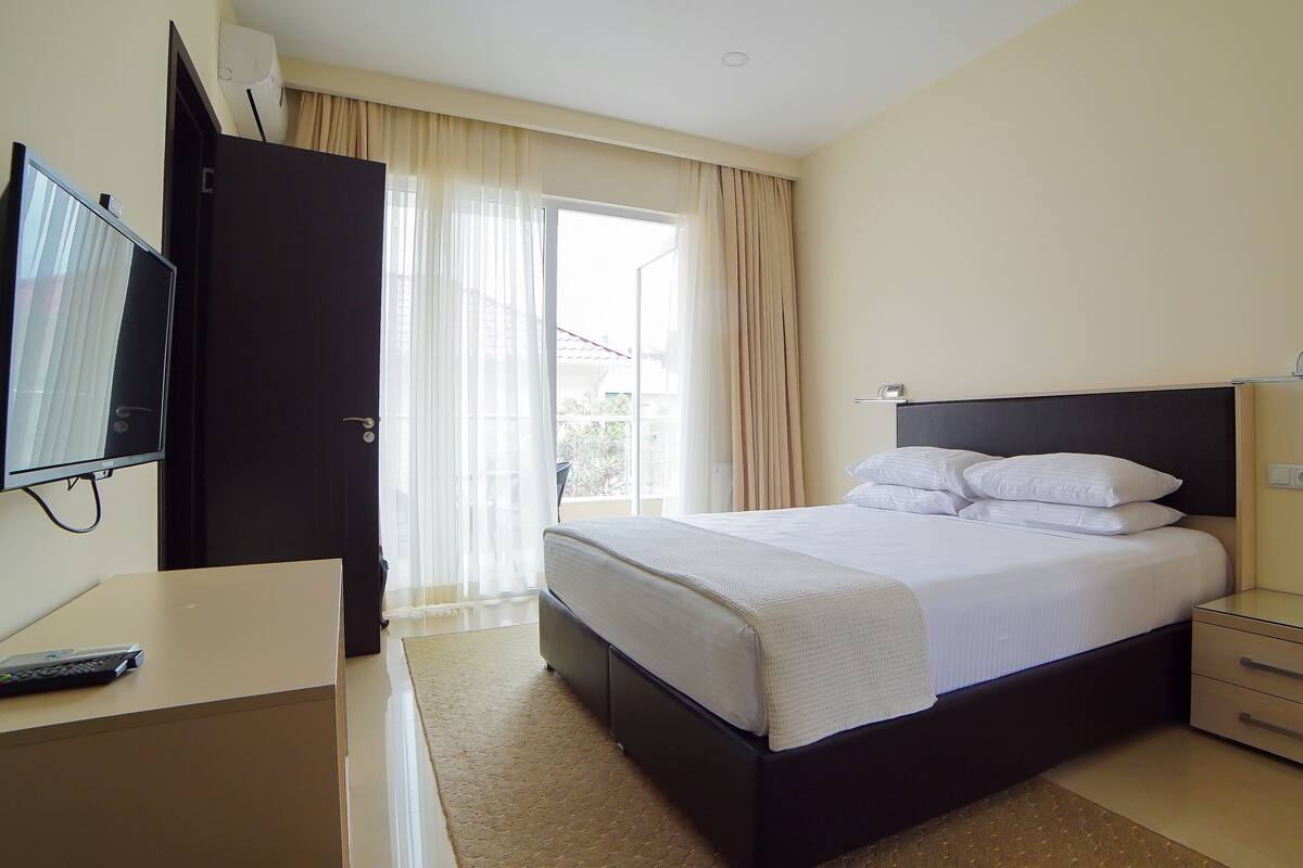 A hotel room with a bed, bedside table, TV, and a view of the outdoors.