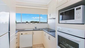 A compact kitchen with white cabinets, a built-in microwave, and a window with a view of the sky and distant landscape. - Ocean Plaza 1466 - Coolangatta Beachfront - Min. 3 Night Stays! (Coolangatta)