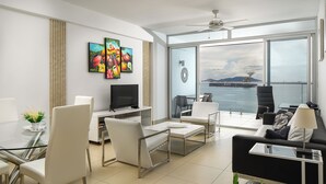 A modern living room with a glass table, white chairs, a black sofa, and a TV. There are large windows with a view of the sea and a ship. - PANAMA CANAL & PANAMA CITY - FULL OCEANFRONT APARTMENT (Panamá City)