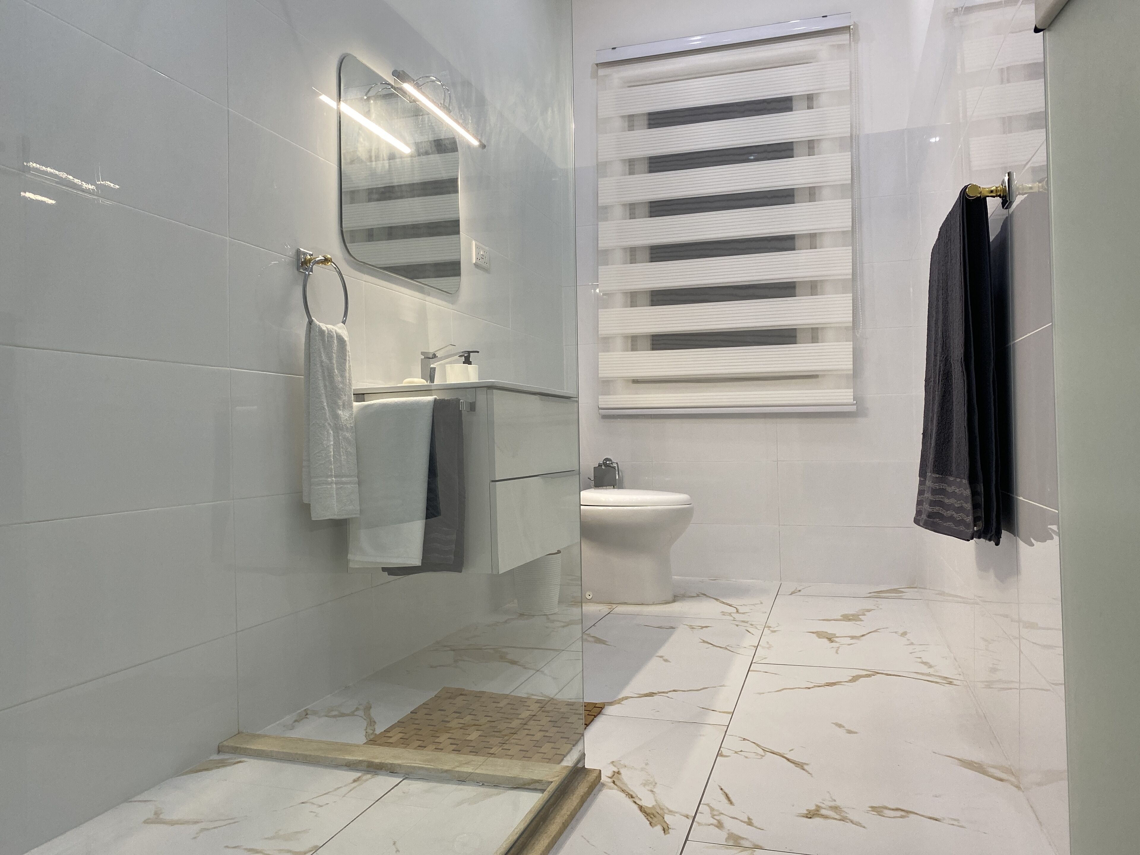 Et moderne badeværelse med hvide fliser, en glasbrusekabine, toilet og et vindue med persienner.