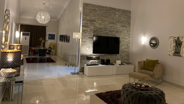 A modern living room with a flat-screen TV, a stone accent wall, a chandelier, and a cozy seating area. - Luxuriously furnished modern home with open-plan design. Tema Spintex (Tema)