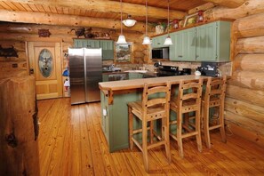 A log cabin kitchen with wooden floors, a central island with bar stools, green cabinets, stainless steel appliances, and pendant lights.