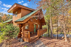 A log cabin with a porch and a green roof, surrounded by trees and fallen leaves. - Big Timber: Big Logs, Bigger Memories (Pigeon Forge)