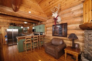 A log cabin interior with a wooden ceiling, a dining area with a bar, a kitchen with a refrigerator, and a fireplace.