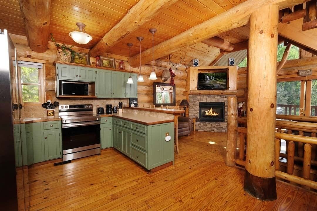 L’intérieur d’une cabane en rondins présente une cuisine avec des meubles verts, une cheminée en pierre et un plancher en bois.