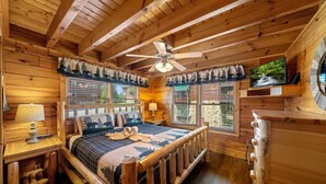 Une chambre dans une cabane en bois, avec un lit, deux tables de chevet, un ventilateur de plafond et un téléviseur posé sur une étagère en bois.