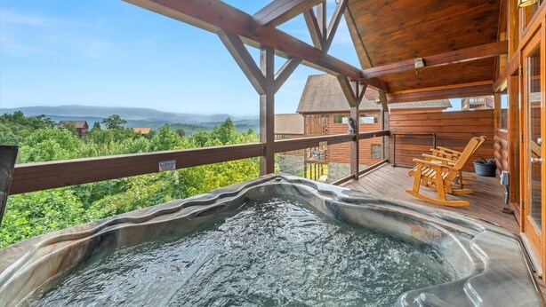 Une cabane en bois dotée d’un bain à remous et de fauteuils à bascule sur une terrasse offrant une vue sur un paysage boisé.