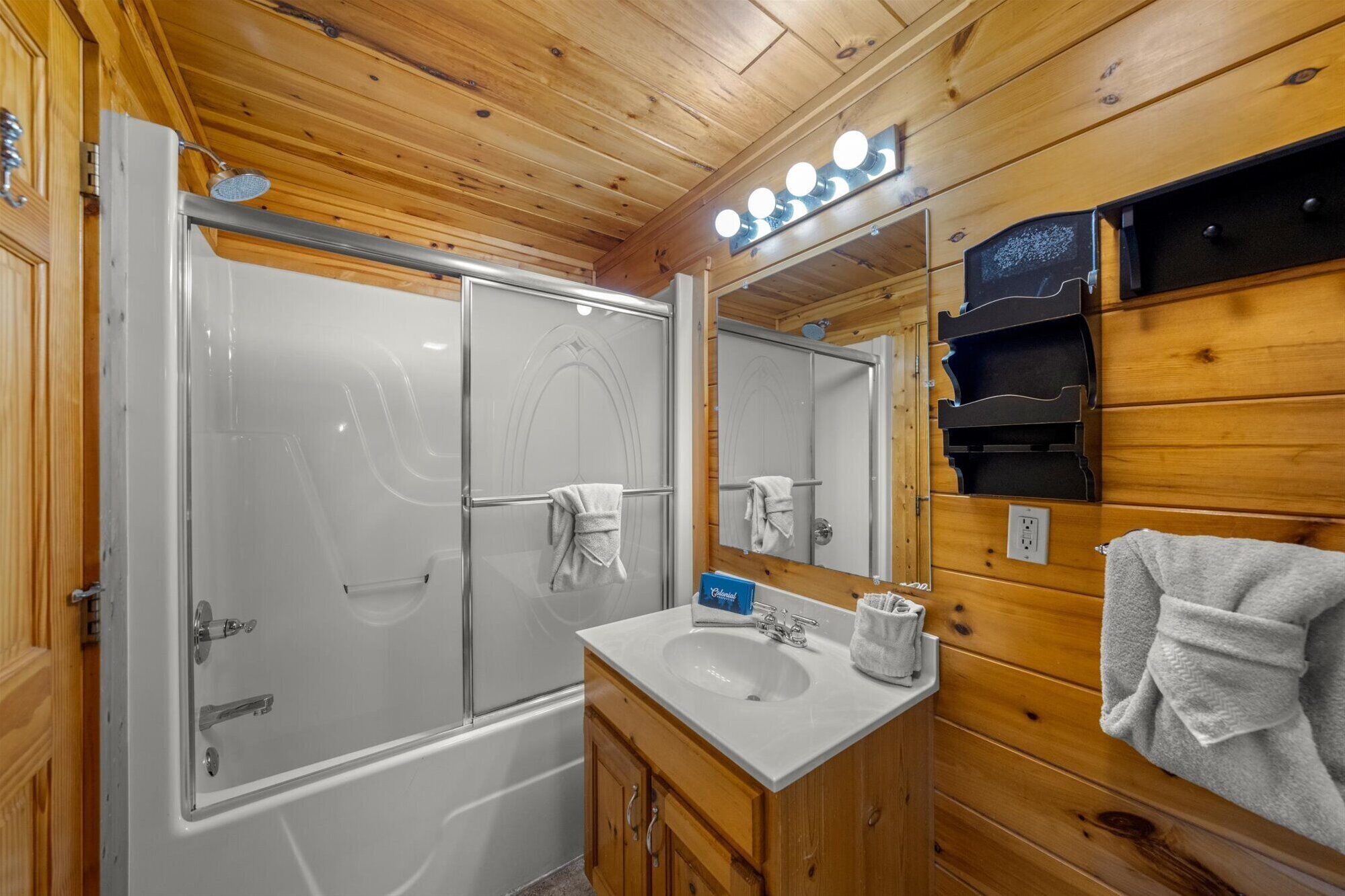 A wooden cabin bathroom with a glass-enclosed shower, a vanity with a sink, and a mirror with lights.