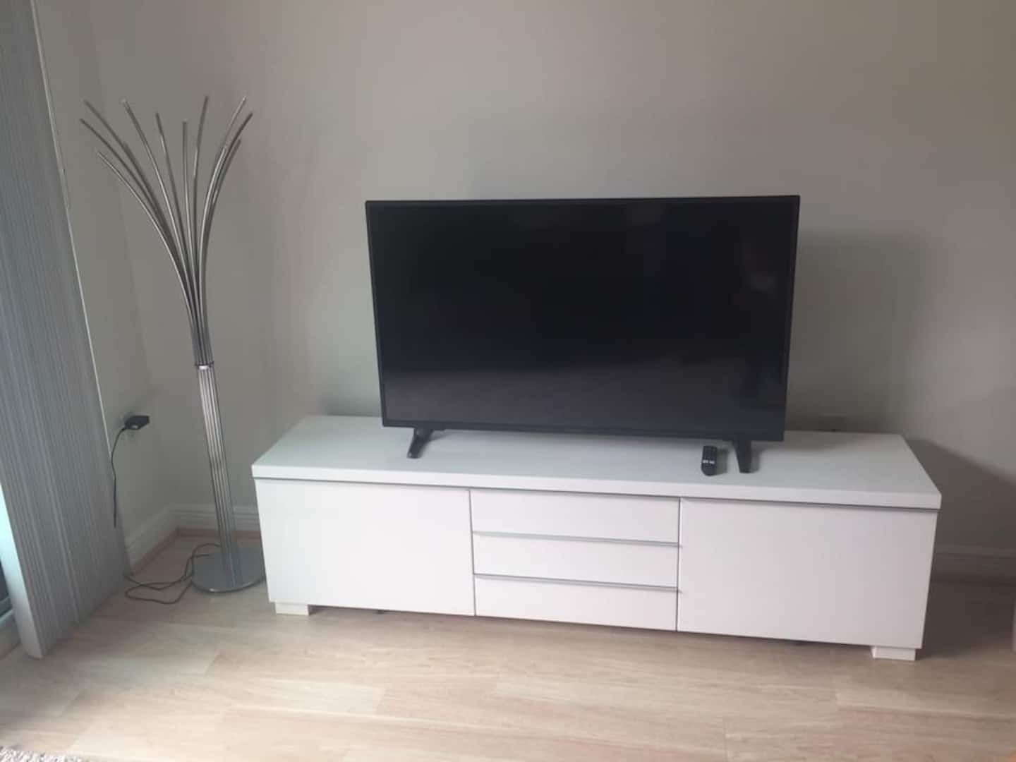 Un televisor de pantalla plana sobre un mueble blanco con cajones, una lámpara de pie y un enchufe en la pared.