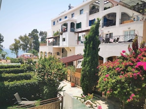 Un edificio de varios pisos con balcones, un jardín con setos y plantas florales, y una vista despejada al mar.