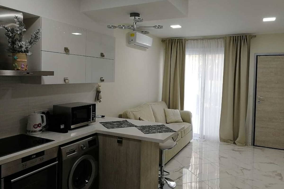 A modern kitchen with a microwave, stove, and dishwasher, a small dining area with a table and chairs, and a living room with a sofa and curtains.