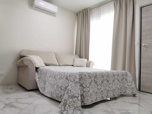 Un dormitorio con cama, sofá, ventana con cortinas y unidad de aire acondicionado.