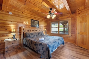 Quarto de cabana de toras com uma cama, uma mesa de cabeceira, um ventilador de teto e uma janela com persianas.