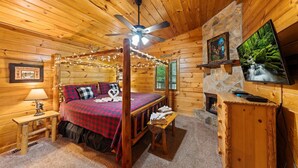 A cozy log cabin bedroom with a four-poster bed, a stone fireplace, a television, and a ceiling fan. - Adjust Your Altitude: Elevate Your Escape (Sevierville)