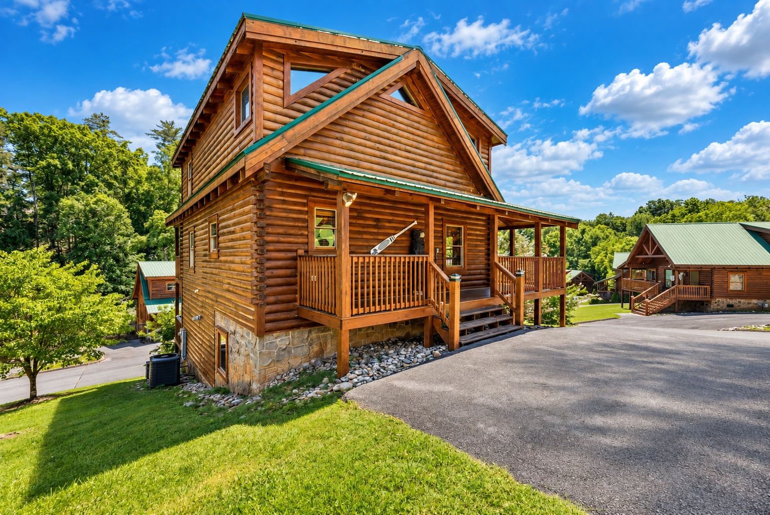 A log cabin with a porch, stone foundation, and a green roof.