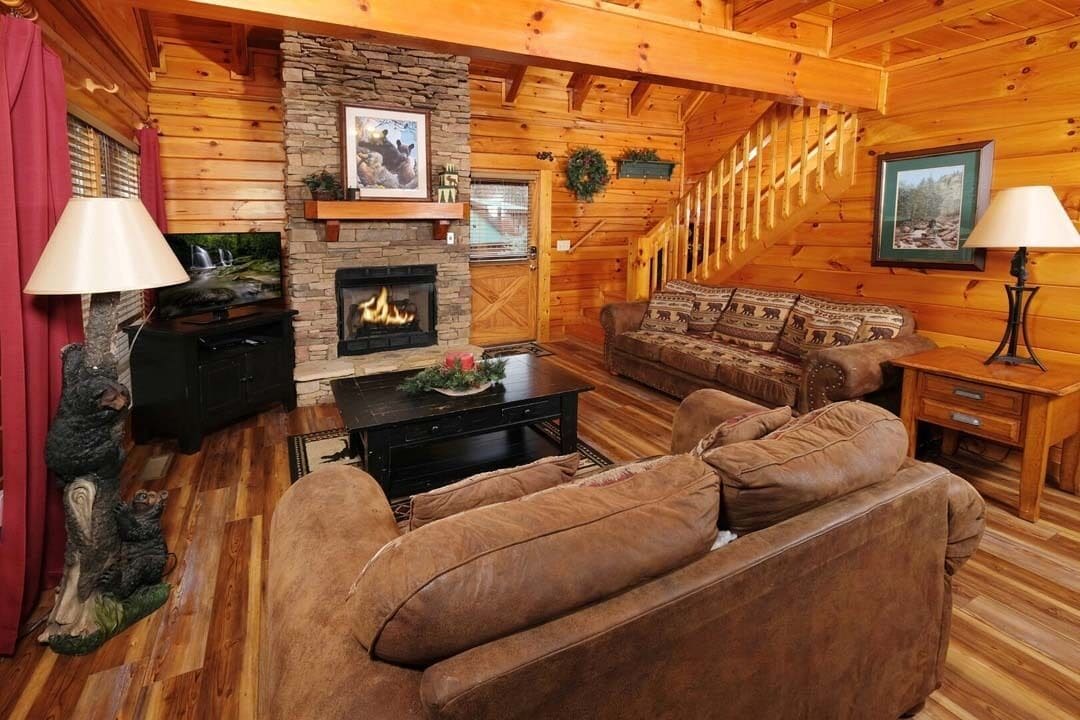 A cozy log cabin interior with a stone fireplace, wooden walls, a leather sofa, and a staircase.