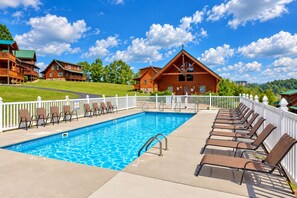 A swimming pool with lounge chairs, a white fence, and wooden cabins in the background.
