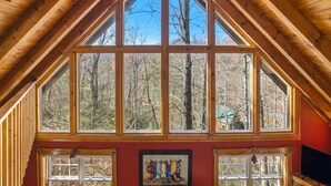 Una stanza con un ampio soffitto di legno a forma triangolare, diverse finestre con vista su una foresta e un quadro appeso al muro.