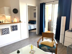 Quarto moderno e bem iluminado, com uma mesa de jantar, uma cadeira, cama com roupa de cama azul, pequena cozinha e vista através de uma porta de vidro.